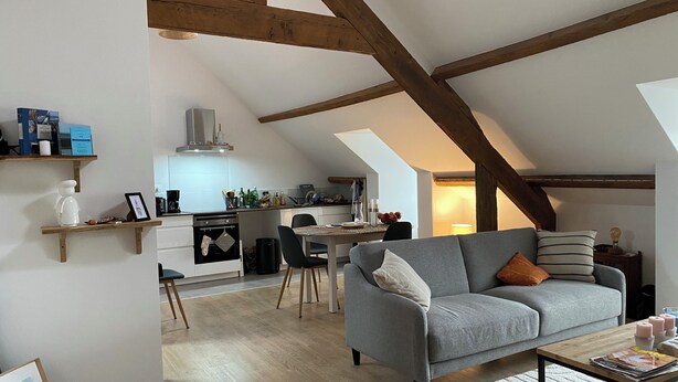 Un espace de vie de style loft avec des poutres apparentes, une cuisine équipée d’appareils modernes, un canapé gris et un coin repas.
