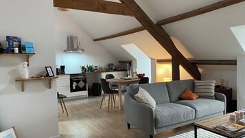 Un espace de vie de style loft avec des poutres apparentes, une cuisine équipée d’appareils modernes, un canapé gris et un coin repas.
