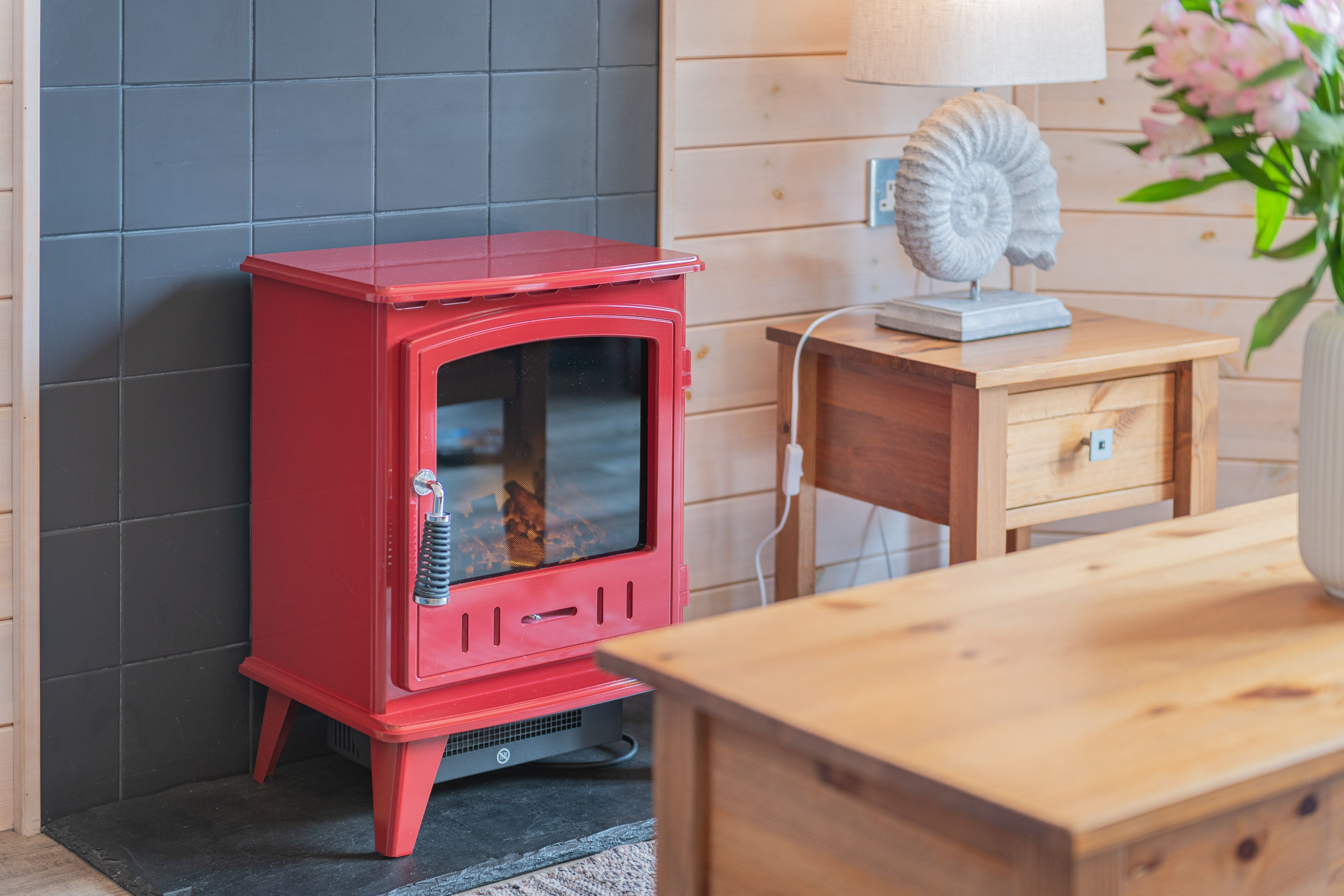 A red wood-burning stove, a wooden table, a lamp, and a flower vase in a room with wooden paneling.