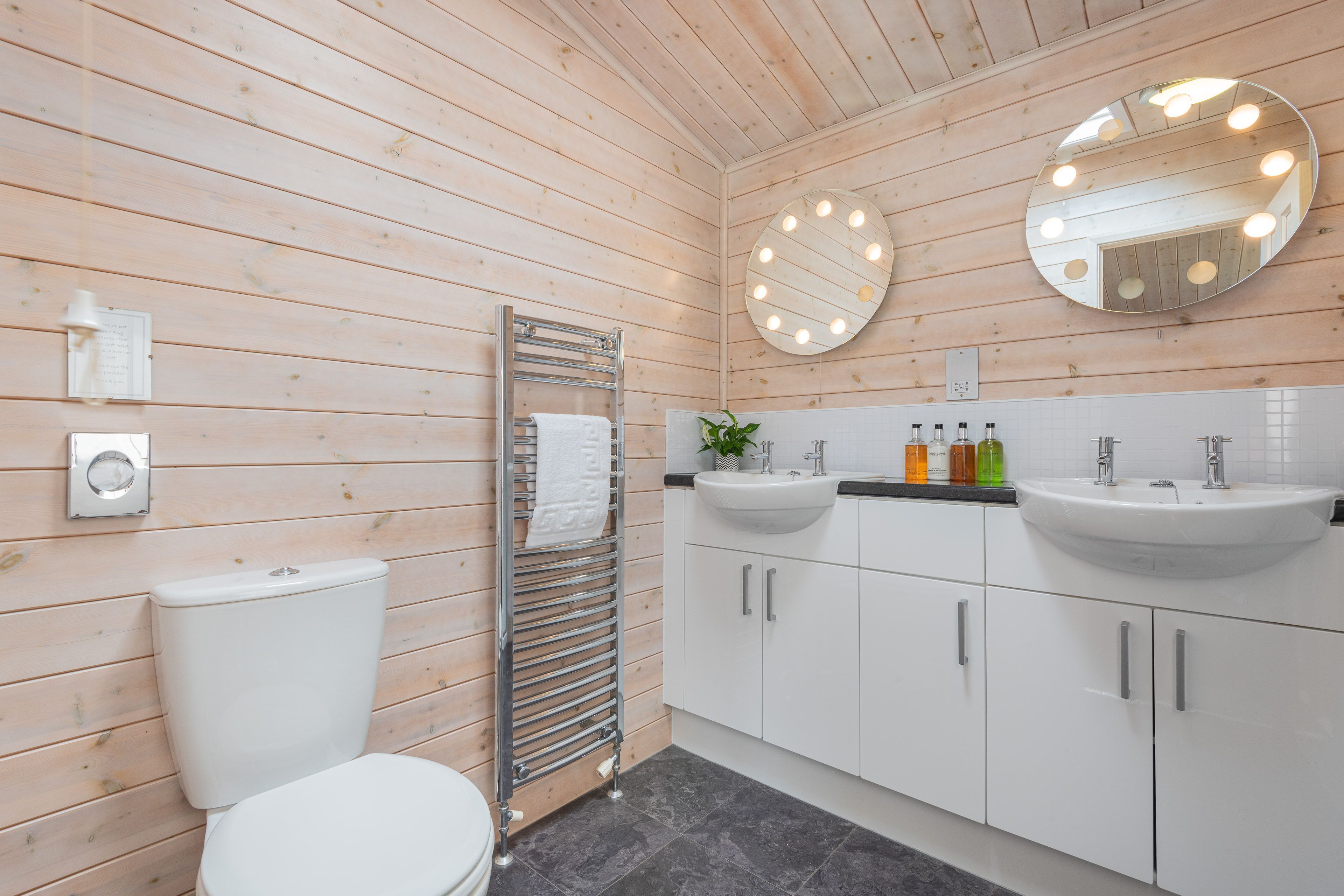 A wooden-paneled bathroom with a toilet, a towel rack, two sinks, and a mirror with lights.