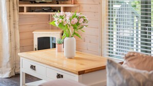 Une table en bois sur laquelle est posé un vase de fleurs, un téléviseur sur une étagère et une fenêtre avec des stores.