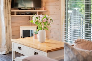 Ein Holztisch mit Blumen in einer Vase, ein Fernseher auf einem Regal und ein Fenster mit Jalousien.