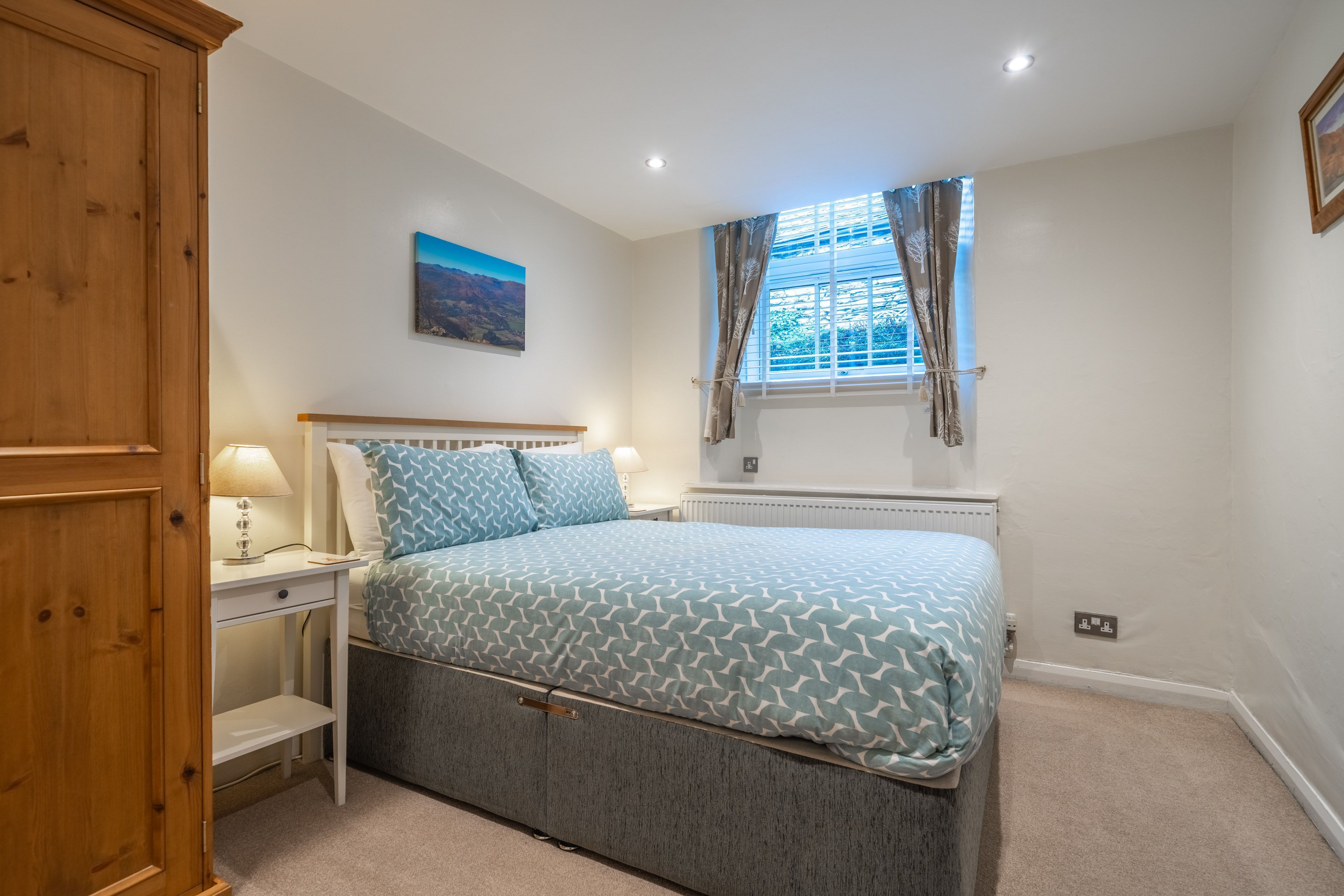 A bedroom with a bed, a nightstand, a lamp, a window with curtains, a radiator, and a wall-mounted picture.
