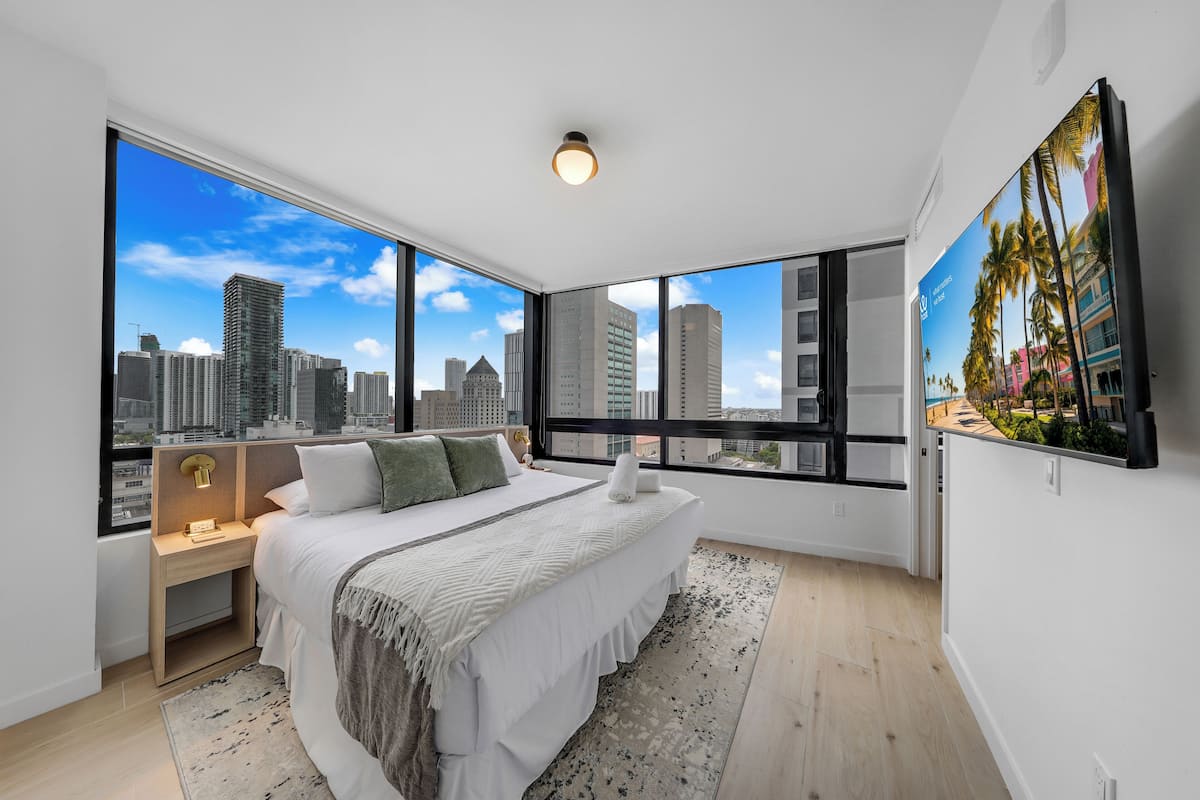 A modern bedroom with a large bed, a nightstand, a wall-mounted TV, and a city view.