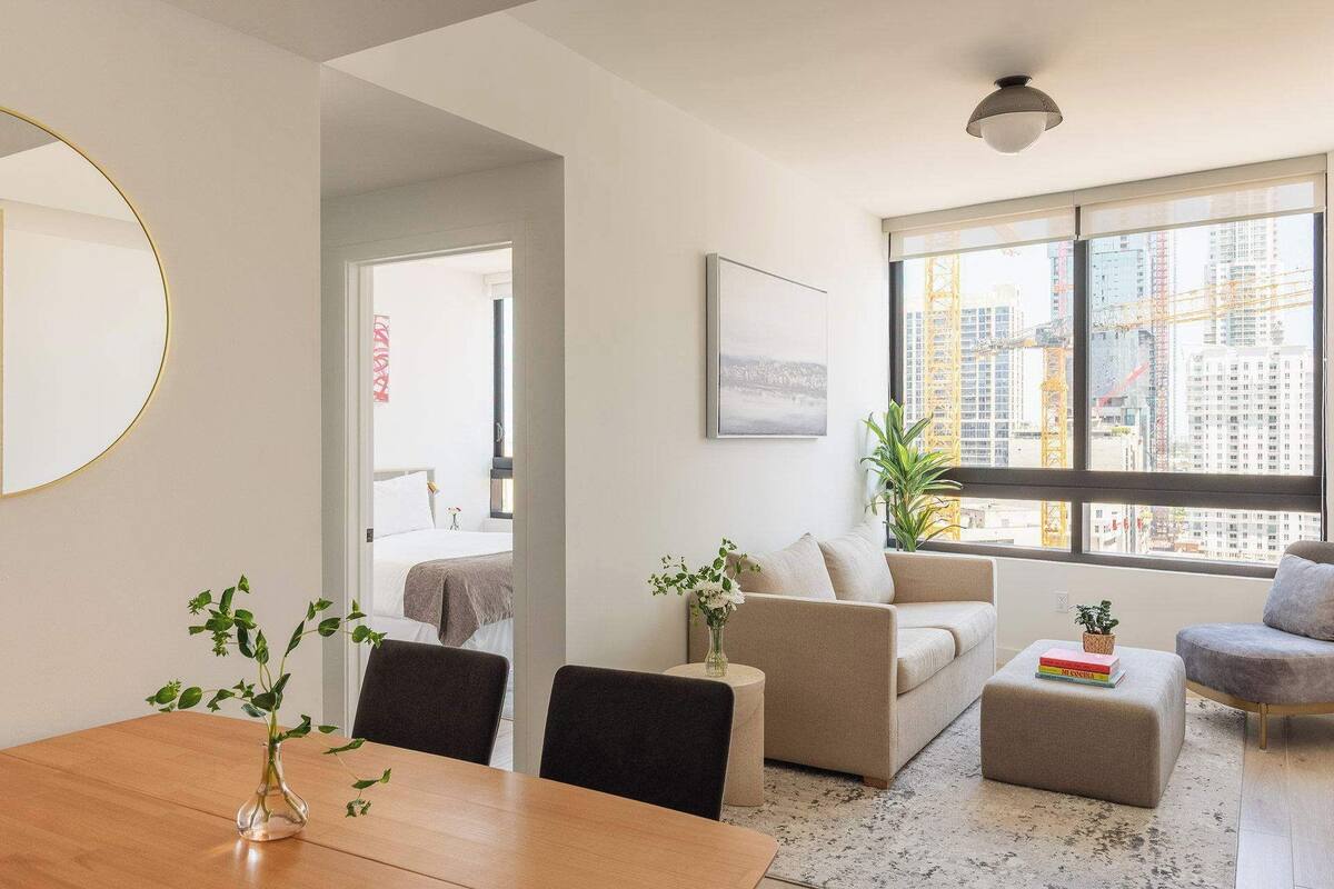 A modern living room with a dining table, sofas, a coffee table, a large window with a city view, and a mirror on the wall.