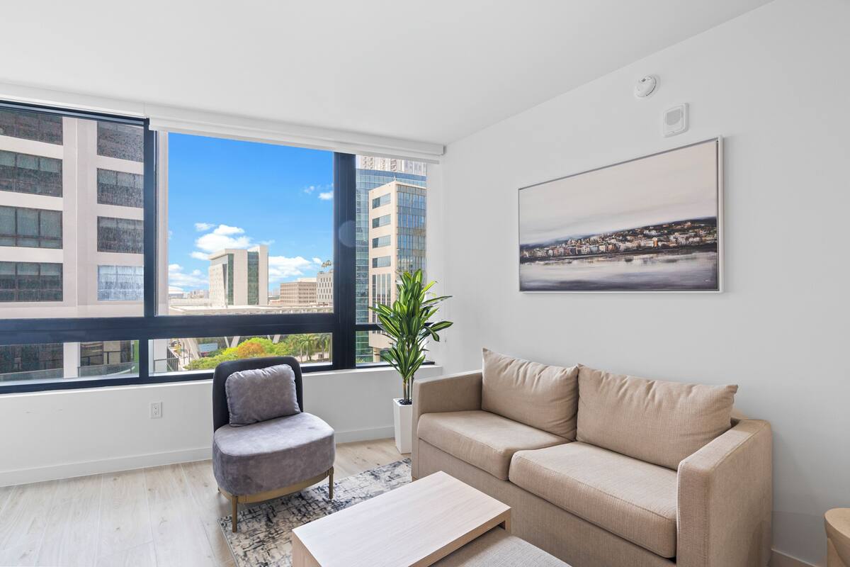 A modern living room with a sofa, armchair, and a large window offering a city view.