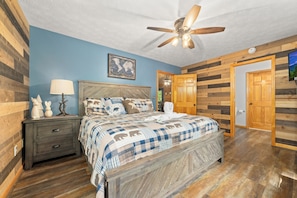 A bedroom with a wooden headboard, a ceiling fan, a bed with a checkered blanket, a nightstand with a lamp, and a wooden door. - Log Cabin, Dog Friendly, Minutes to Attractions, Hot tub, Fire-pit, Peaceful! (Sevierville)