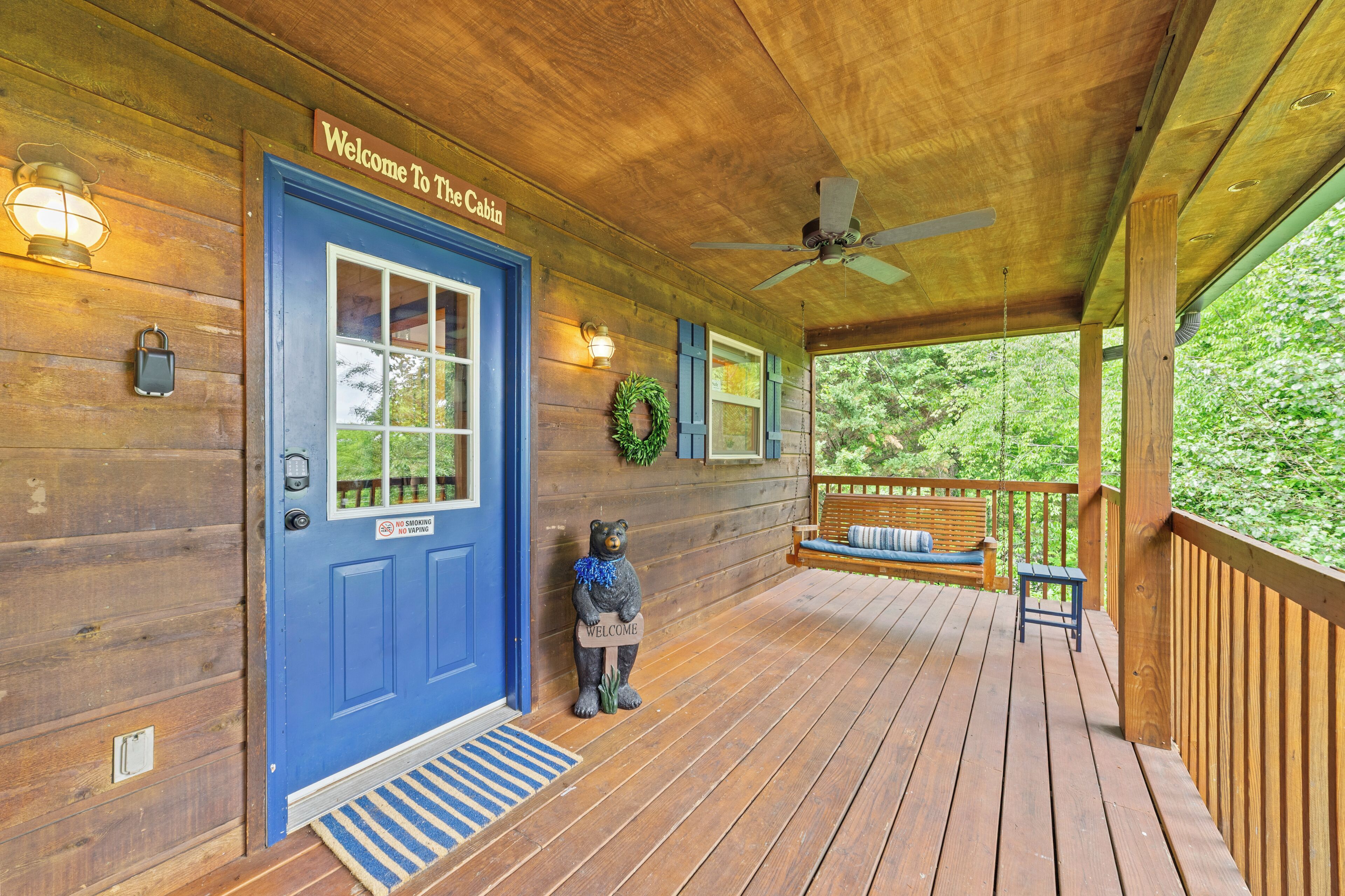 Une cabane en bois avec une porte bleue, une pancarte de bienvenue, une statue d’ours, un banc et un ventilateur de plafond.