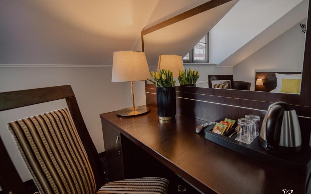 Habitación de hotel con un escritorio de madera, una lámpara, una planta en maceta, una tetera y una bandeja con un libro y vasos.