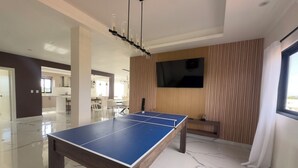 Ett bordtennisbord i ett modernt rum med en TV, en väggpanel i trä och utsikt över utomhusmiljön.