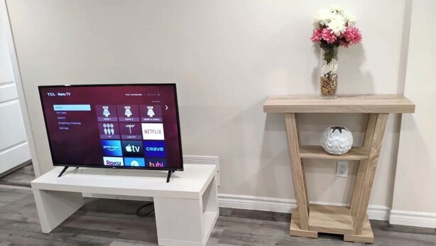 A television on a white stand, a wooden shelf with a vase and a decorative item, and a wall with a power outlet. - Fully Private Unit in Heart of Scarborough with Private Kitchen (Toronto)