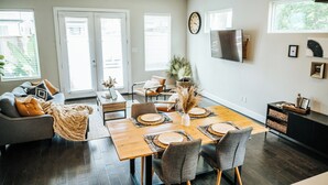 A dining area with a wooden table set for four, surrounded by grey chairs, a grey sofa with orange cushions, and a black cabinet with baskets. - Stay in the Heart of Houston, 5' to Downtown, close Medical Center & World Cup (Houston)