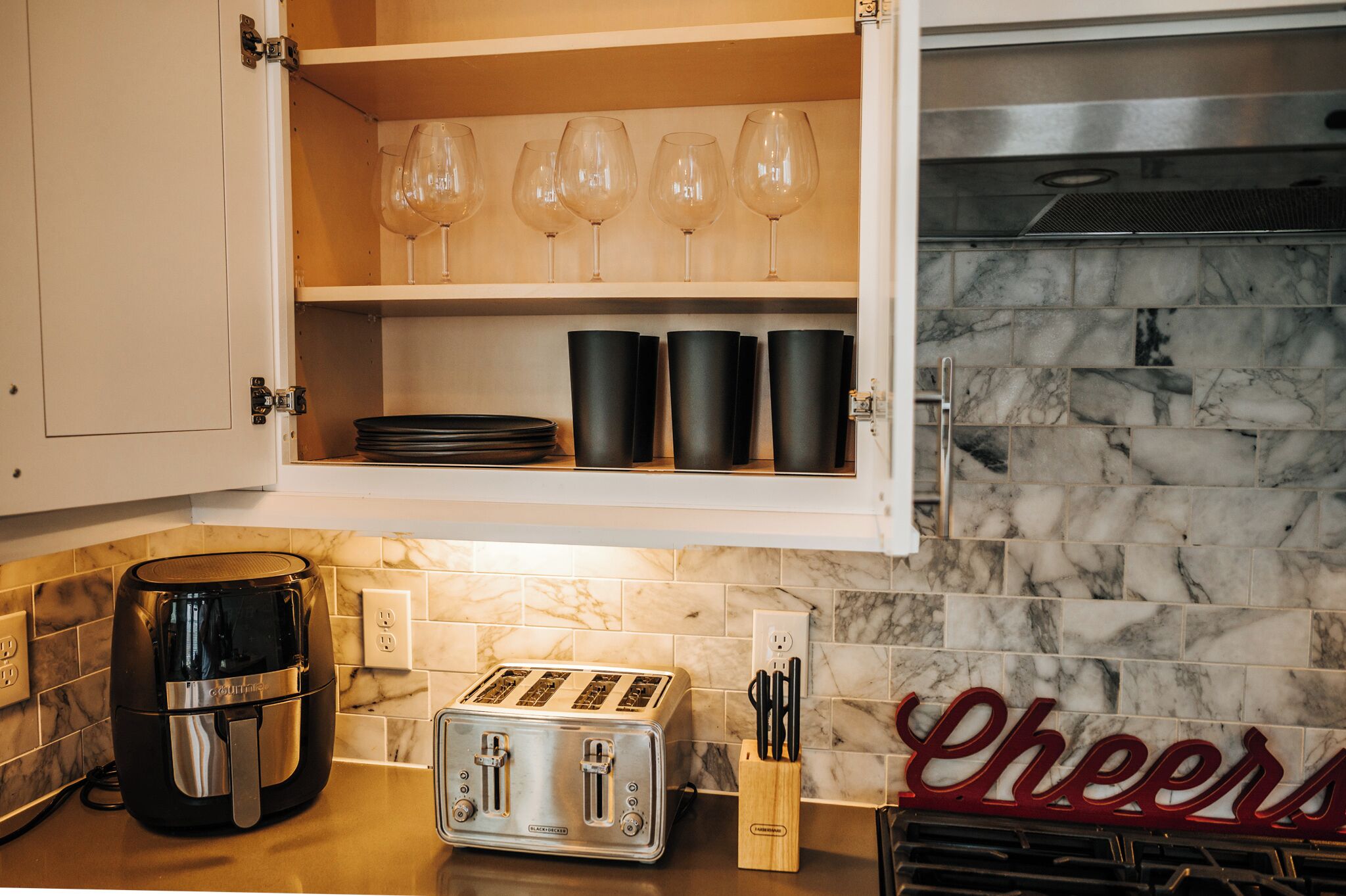 A kitchen with a built-in microwave, toaster, and a shelf with wine glasses and black cups.