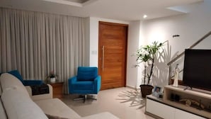 Un salon moderne avec un canapé, un fauteuil bleu, un téléviseur, une plante en pot et une porte en bois.