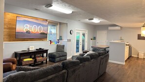 Ein Wohnzimmer mit Ecksofa, einem an einer Holzwand montierten Fernseher, einem Fenster mit Aussicht und einer zur Terrasse führenden Tür.