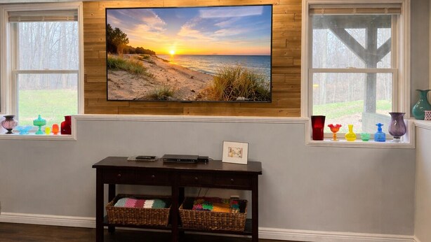 Ein Wohnzimmer mit an einer Holzwand montiertem Fernseher, einem dunklen Holzkonsole mit Körben und Fenstern mit dekorativen Vasen.