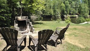 En eldstad omgiven av Adirondack-stolar i ett skogsområde med en sjö i närheten.