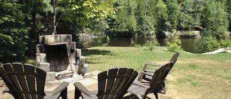 A fire pit surrounded by Adirondack chairs in a wooded area with a lake.