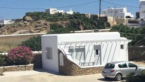 A white building with a sloped roof, a car parked in front, and a hillside with vegetation in the background. - Diogenis Village Mykonos (Mykonos)