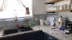 A kitchen with a granite countertop, a sink, a window with a curtain, and a shelf with glassware.