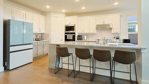 A modern kitchen with white cabinets, a stainless steel refrigerator, a microwave, and a built-in oven. There are four bar stools at the island.