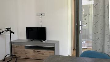 Une chambre moderne avec un téléviseur à écran plat posé sur une console grise, un meuble en bois, un fauteuil de bureau noir et une porte en verre donnant sur un balcon.