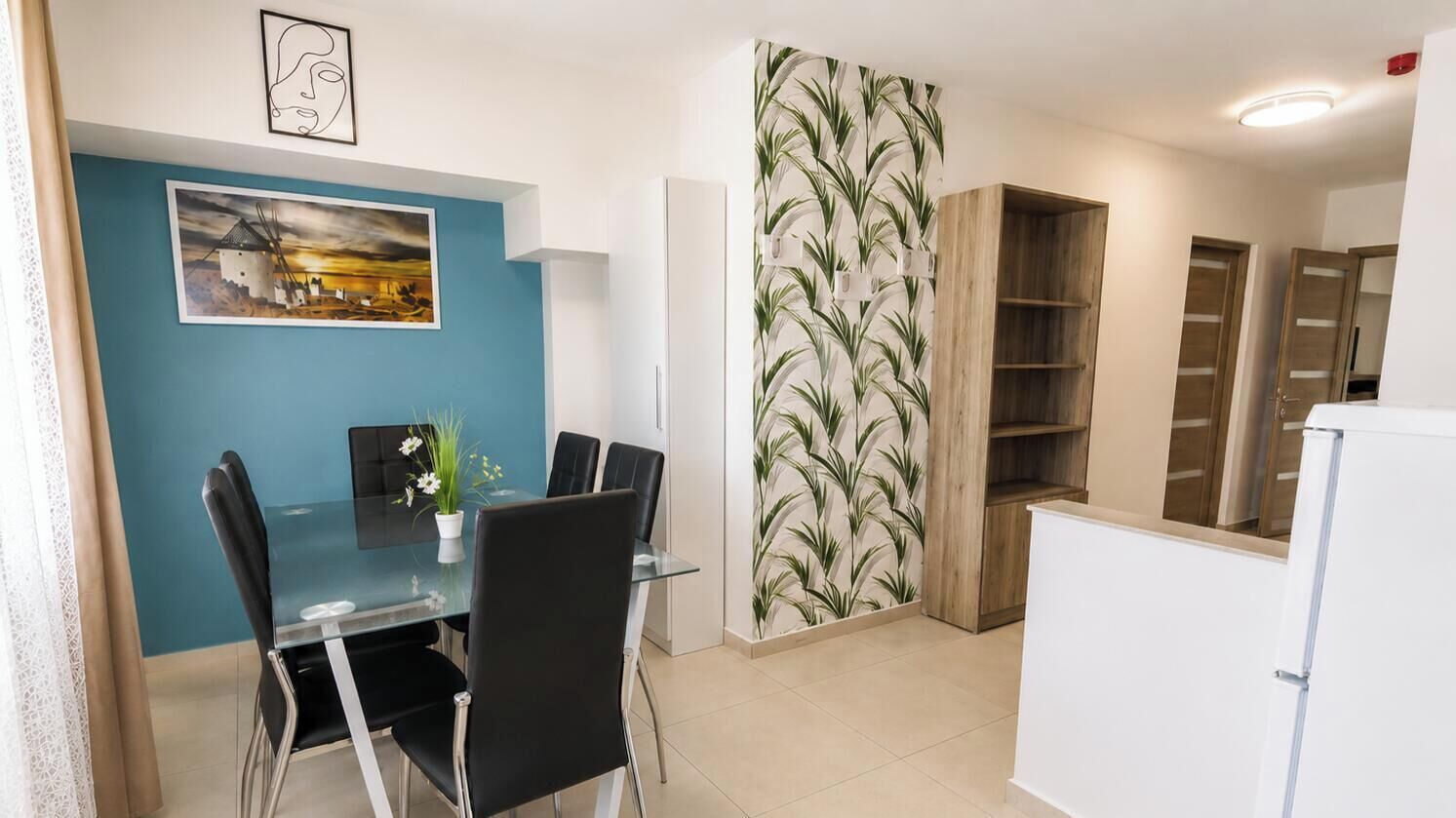 A dining area with a glass table and chairs, a painting on the wall, and a wooden bookshelf.