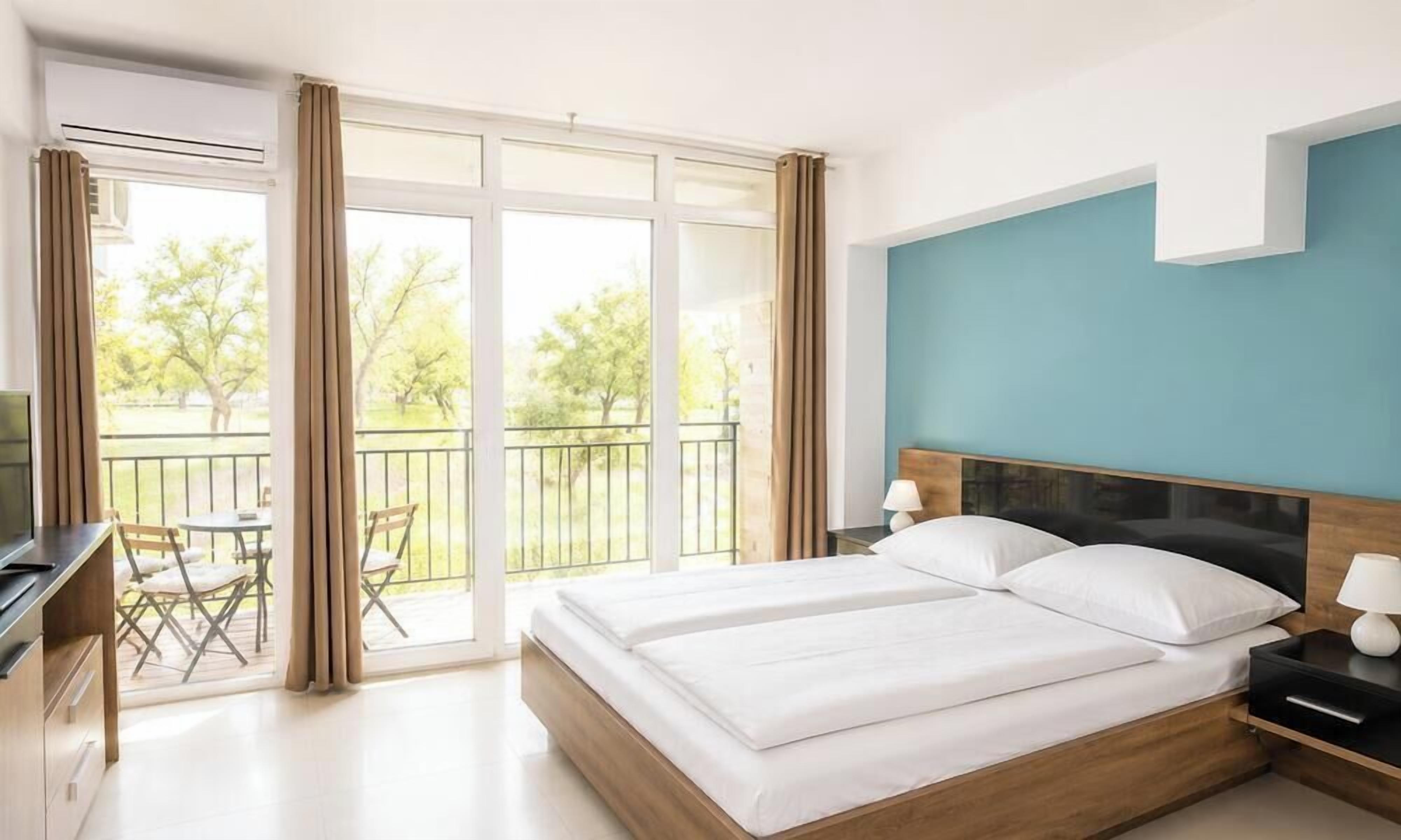 A hotel room with a large bed, a desk, a chair, and a balcony with a view of trees.