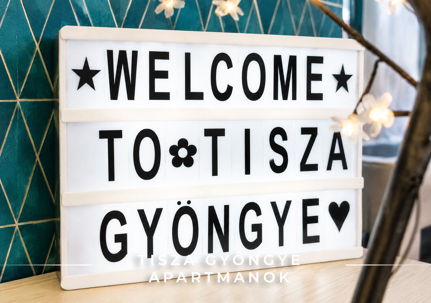 Una caja de luz con el texto 'WELCOME TO TISZA GYÖNGYE APARTMANOK' en letras negras sobre un fondo blanco.