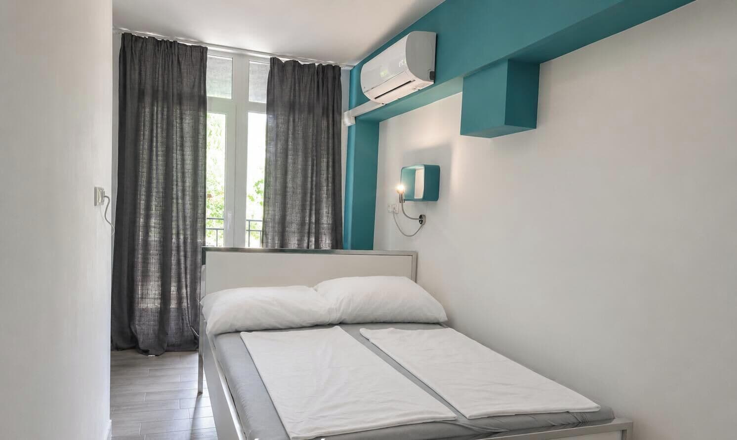 Una cama individual con sábanas blancas en una habitación con detalles en color turquesa, una ventana con cortinas y un equipo de aire acondicionado.