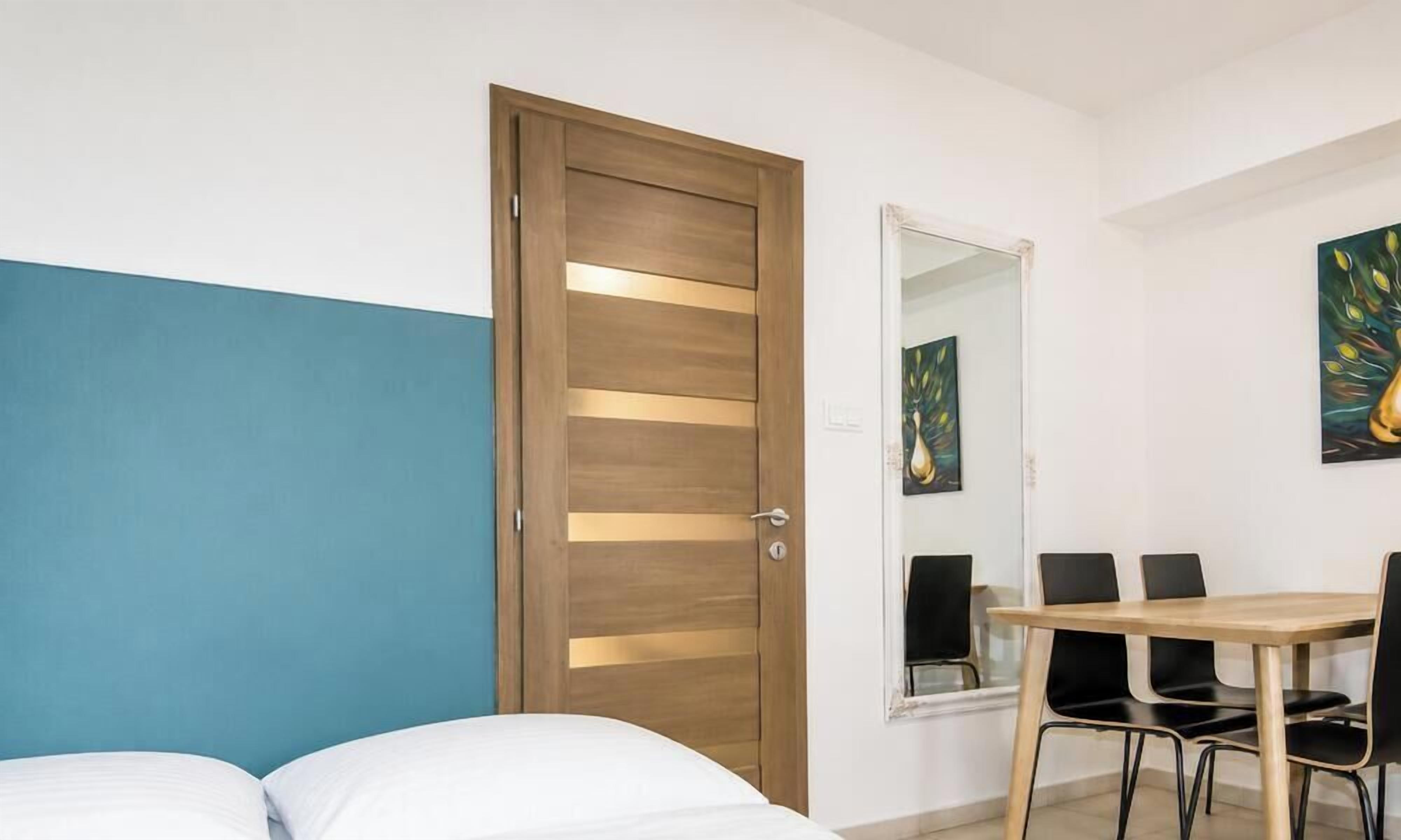 A bedroom with a bed, a wooden door, a dining table with chairs, and a mirror.
