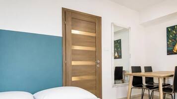 A bedroom with a bed, a wooden door, a dining table with chairs, and a mirror.