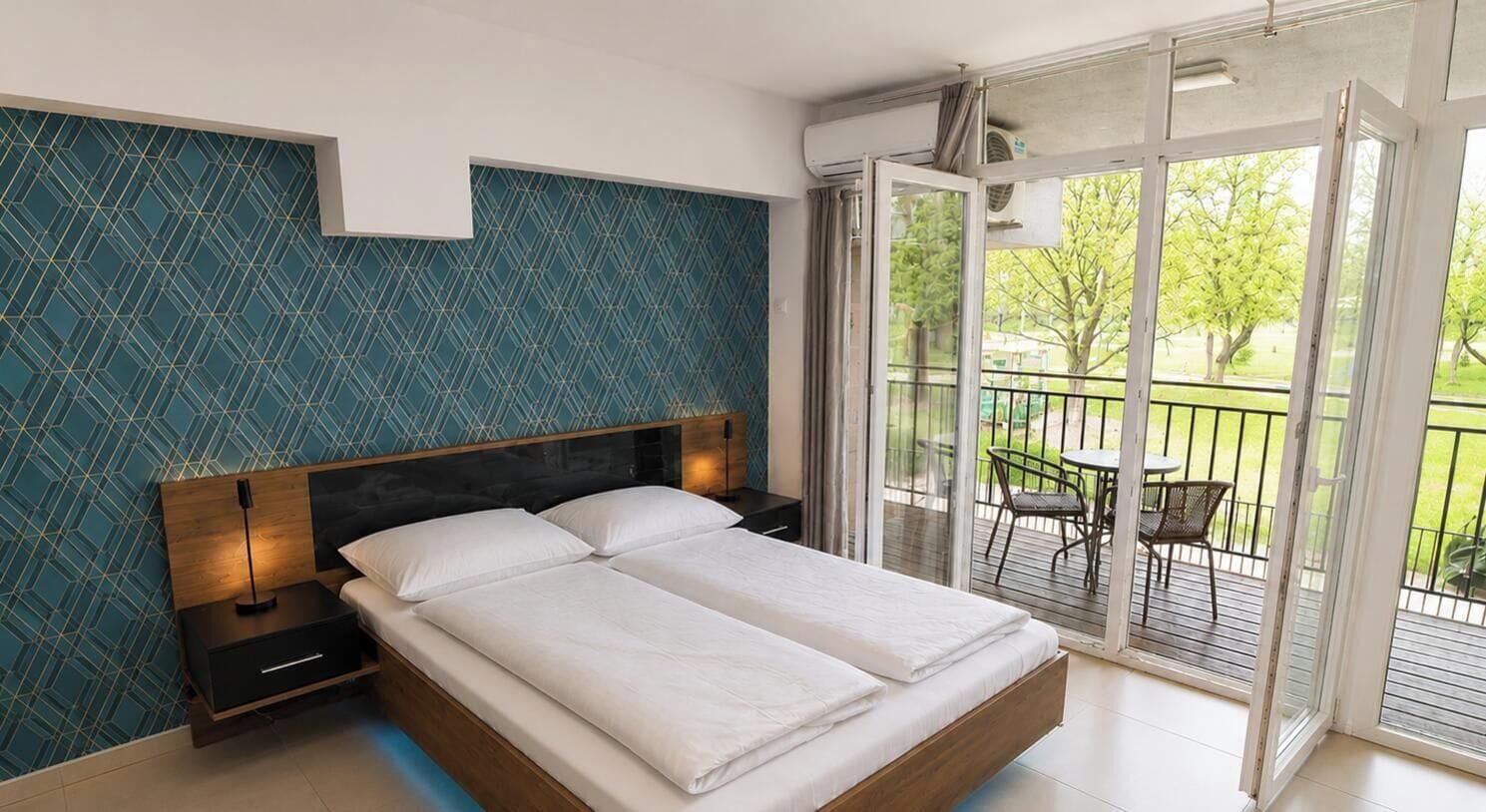 A modern bedroom with a large bed, a nightstand, and a view of a balcony with outdoor furniture.