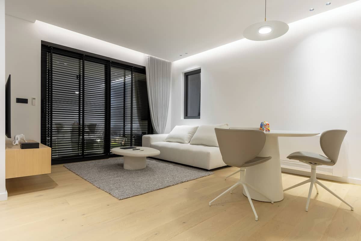 A modern living room with a white sofa, a dining table with chairs, a coffee table, and a large window with blinds.