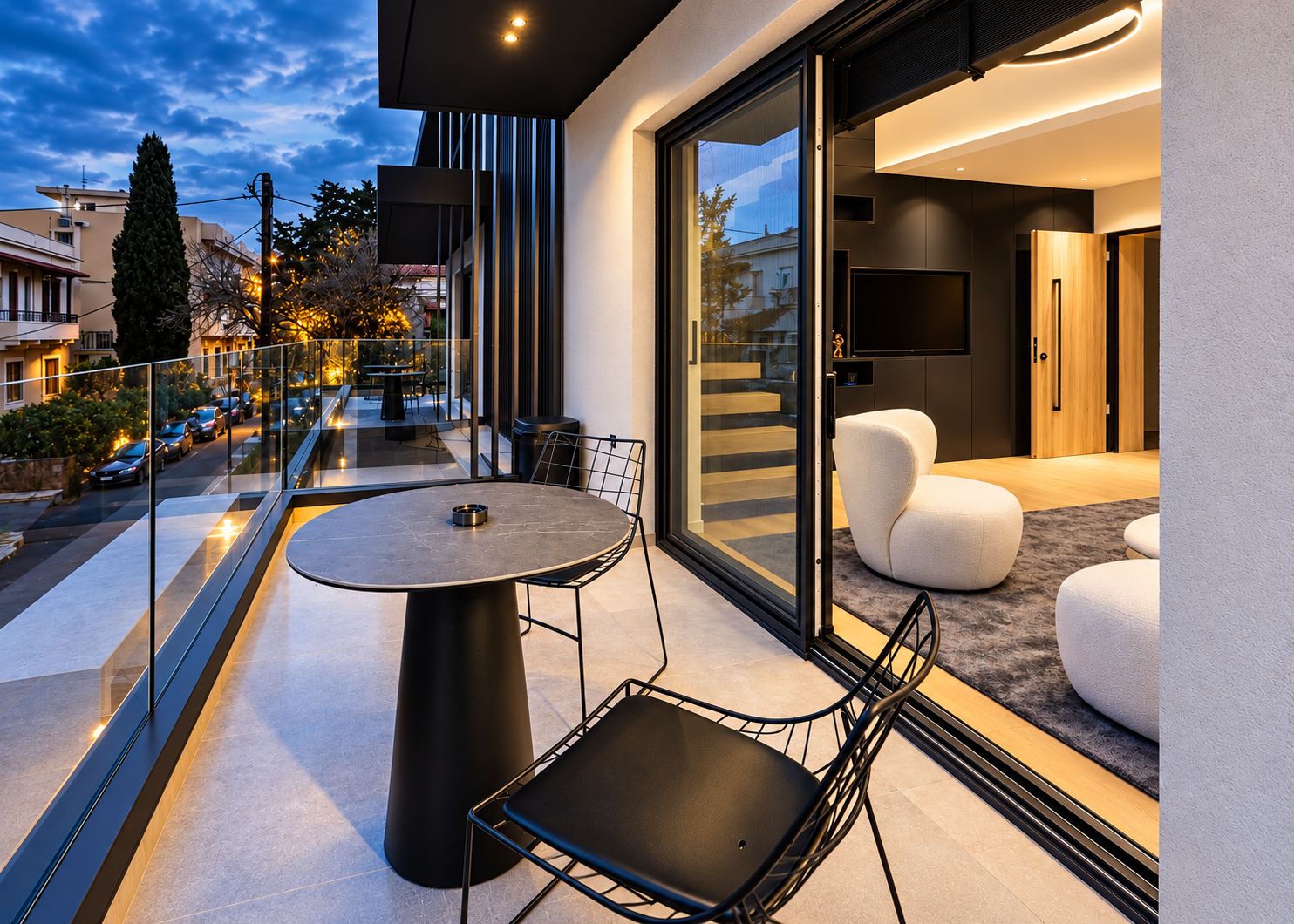 Un balcón moderno con puerta de cristal que da a un espacio interior, una mesa redonda con sillas y vista a la calle.