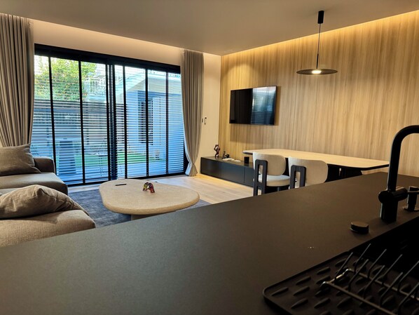 A modern living room with a sofa, a round coffee table, a TV mounted on the wall, and a dining area with chairs. - KALVOKORESI 16 I PREMIUM APARTMENTS (Chios)