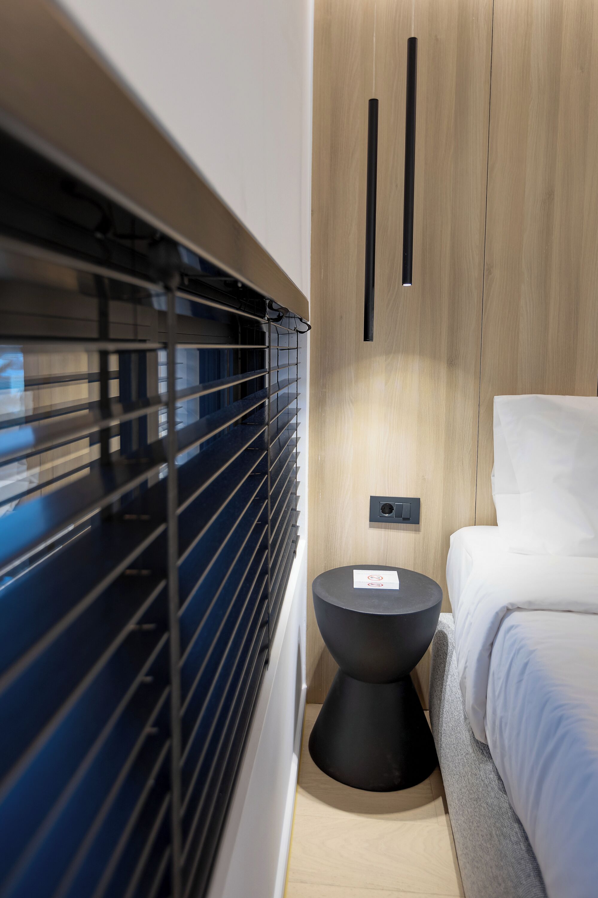 Une chambre moderne avec un lit, une petite table ronde, un panneau de commande fixé au mur et une fenêtre avec des stores.