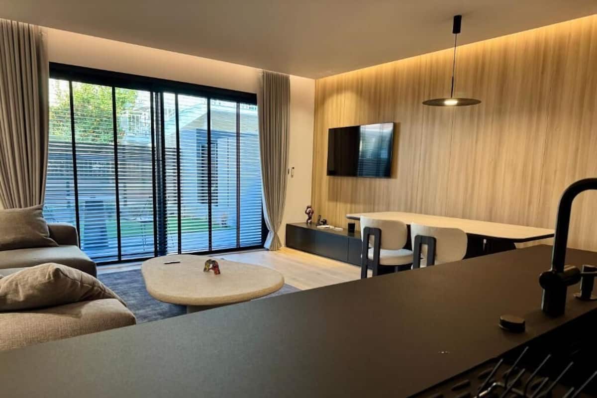A modern living room with a sofa, a round coffee table, a dining area with a table and chairs, and a TV mounted on the wall.