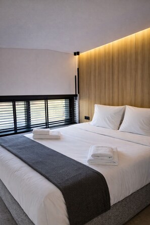 A modern hotel room with a large bed, a wooden headboard, and a view through the window blinds. - KALVOKORESI 16 I PREMIUM APARTMENTS (Chios)