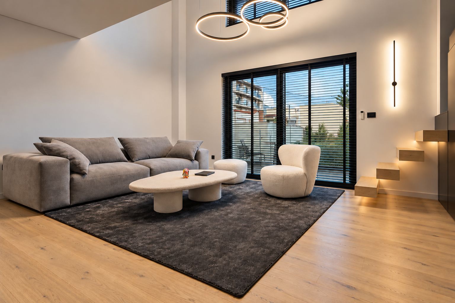 A modern living room with a sofa, armchair, and coffee table, a staircase with built-in lighting, and a large window with blinds.