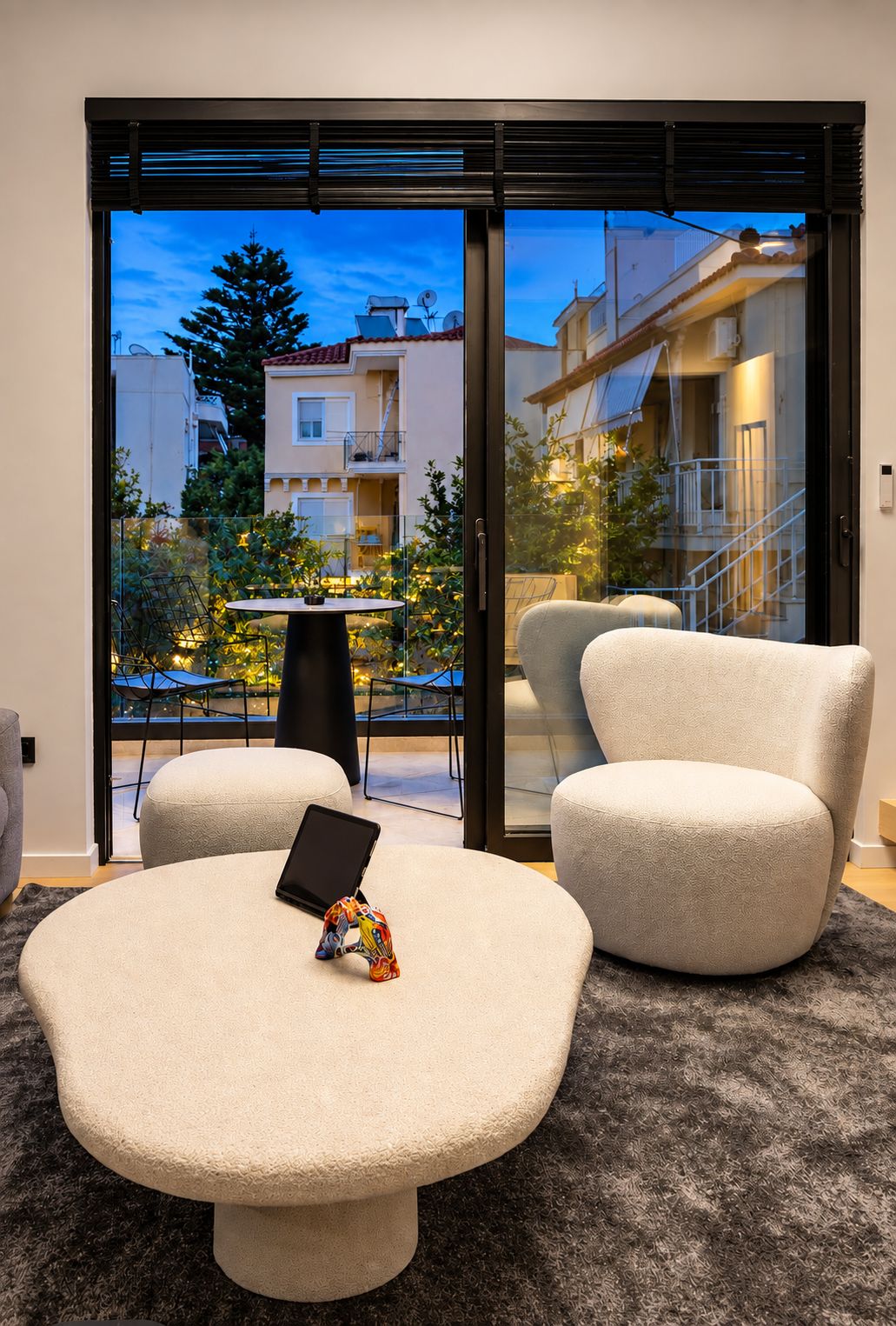 Un salon moderne avec une table basse ronde, deux fauteuils et une porte vitrée donnant sur un balcon extérieur d’où l’on peut admirer les bâtiments et les arbres.