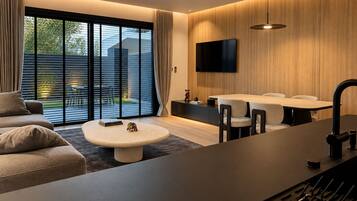 A modern living room with a sofa, a round coffee table, a dining area with chairs, a TV mounted on the wall, and a large window with blinds.