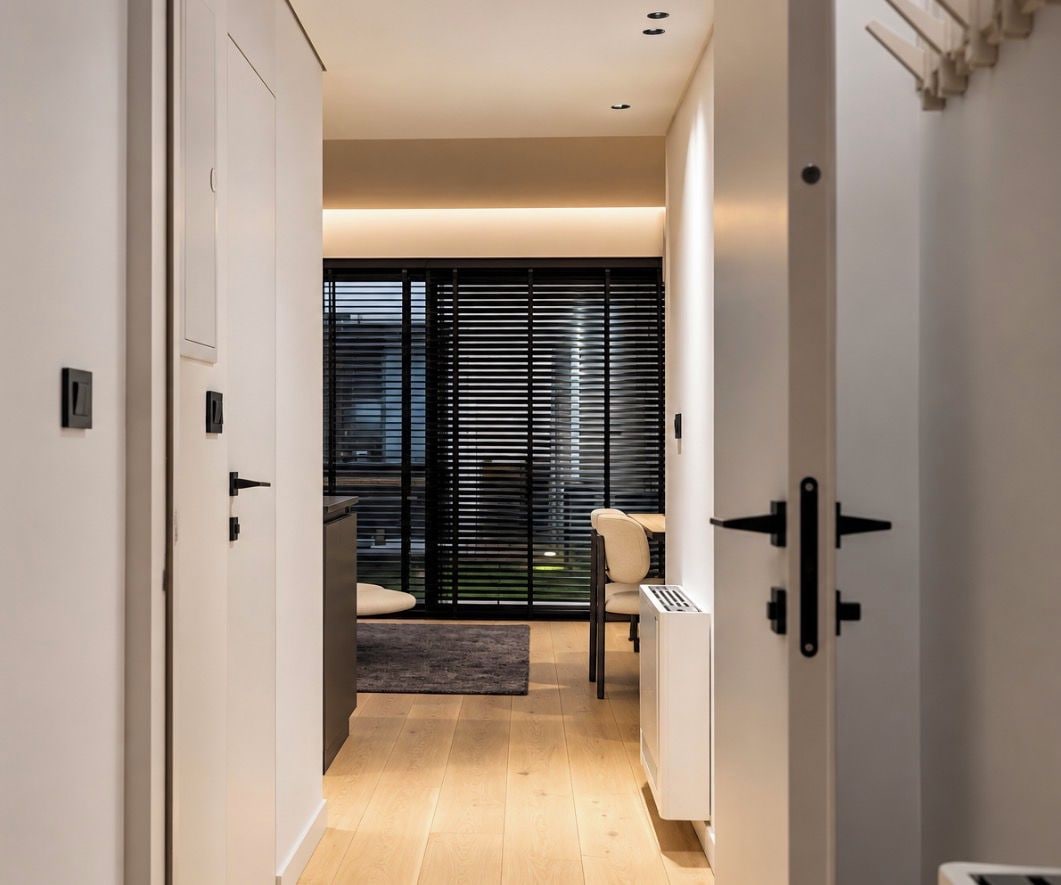 Un pasillo con una puerta que da a una habitación con ventana, un radiador y una silla.