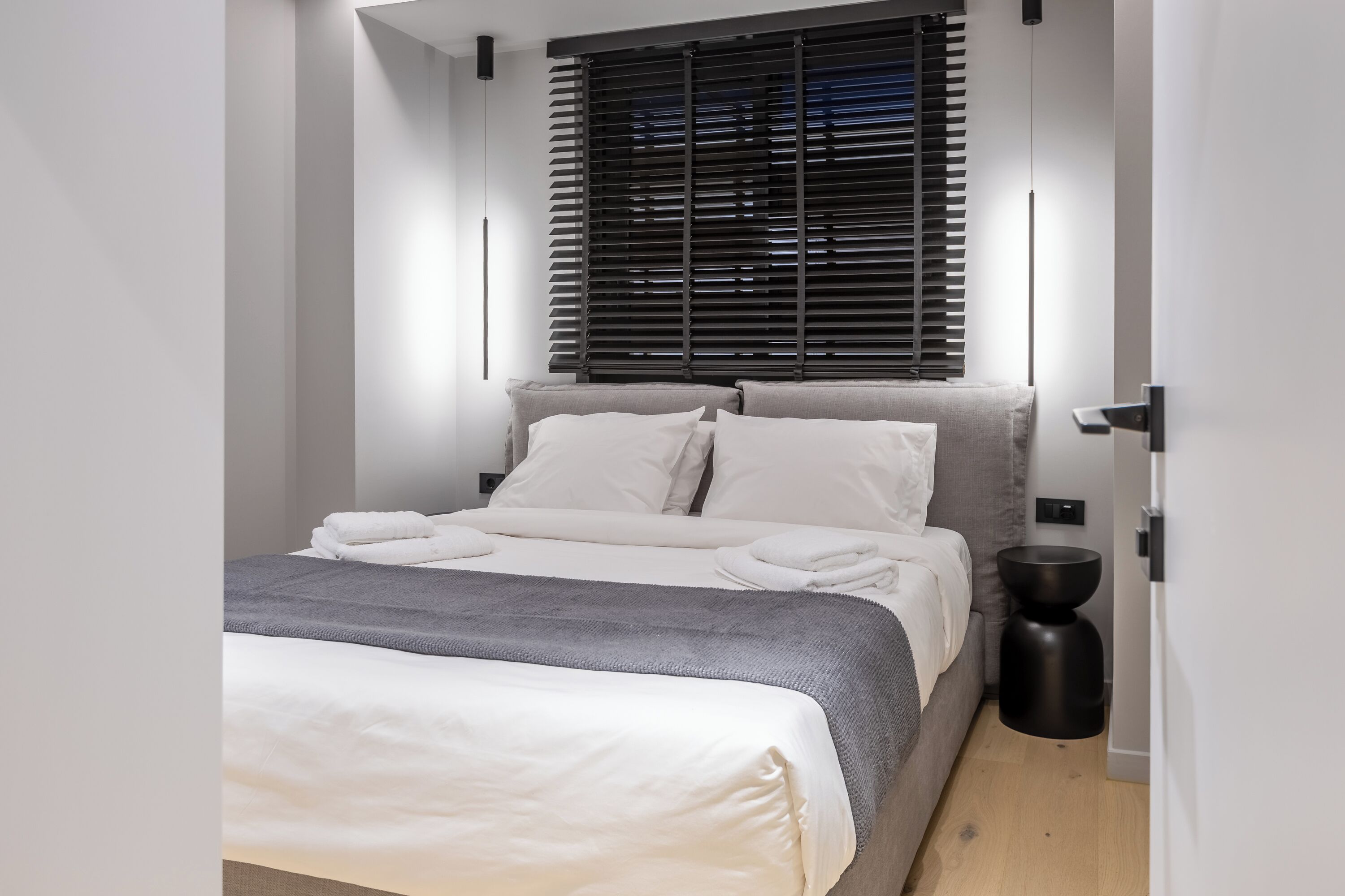 Un dormitorio moderno con una cama, ropa de cama gris y blanca, una mesita de noche negra y una ventana con persianas.