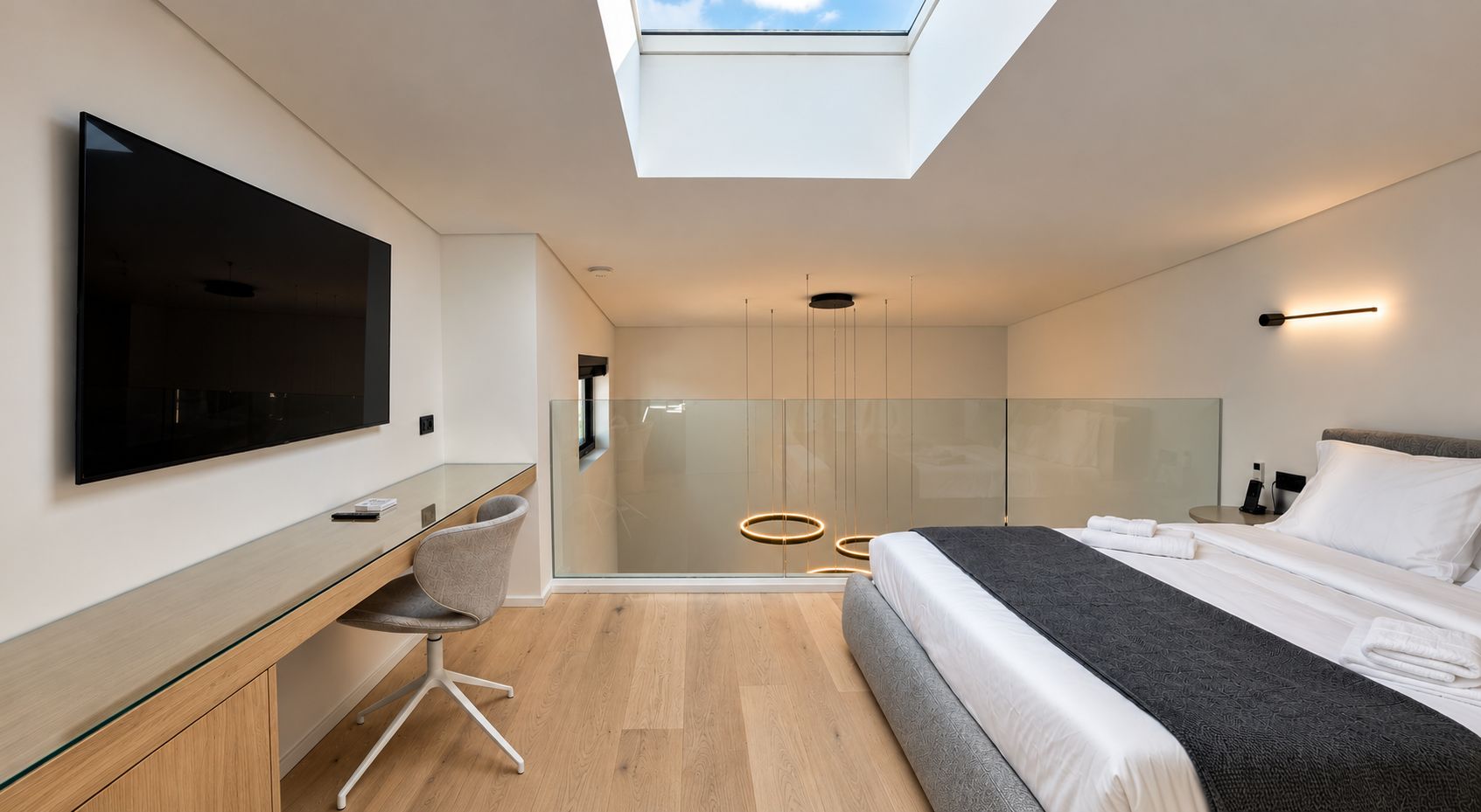 Habitación de hotel moderna con tragaluz, un televisor de pantalla plana grande, un escritorio con silla, cama con cabecera gris y un separador de vidrio.