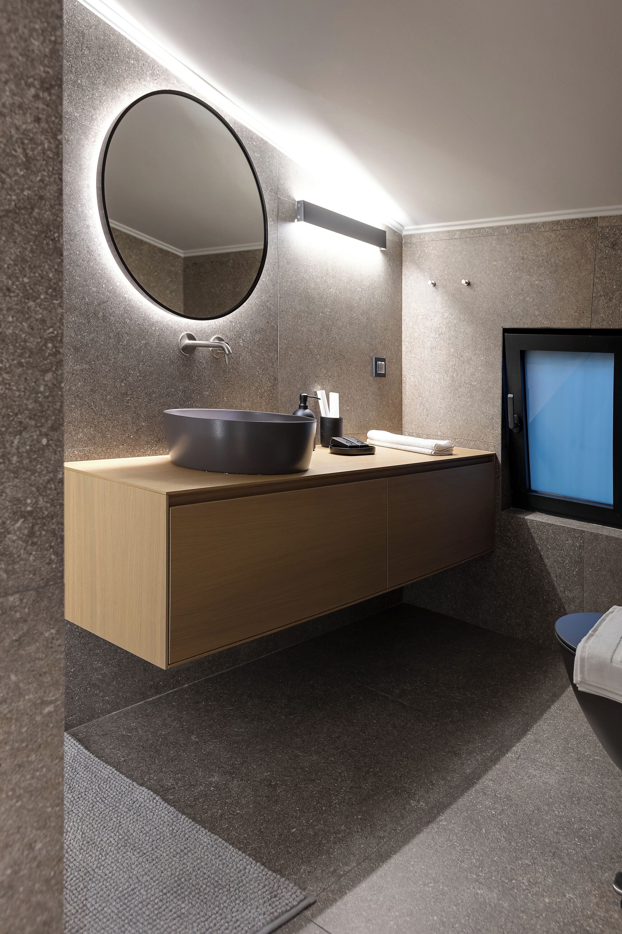 A modern bathroom with a round mirror, a vessel sink, a towel, and a toilet.