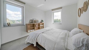 Une chambre à coucher avec un lit, deux fenêtres, une étagère en bois et une vue sur l’extérieur.