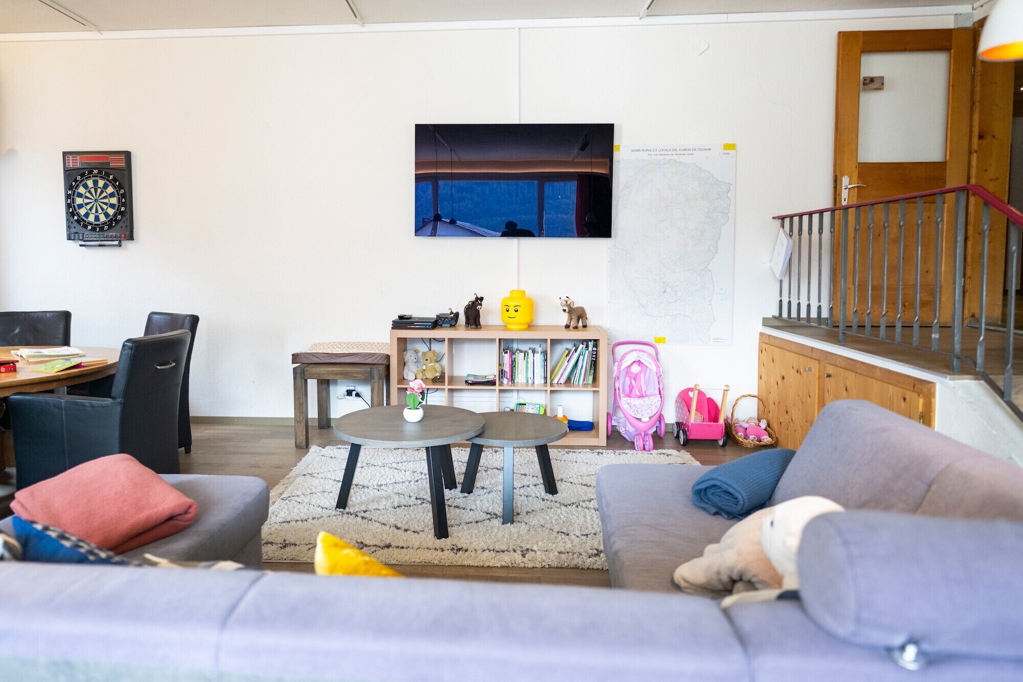 Een woonkamer met een bank, salontafel, televisie aan de muur, boekenkast en een trap.
