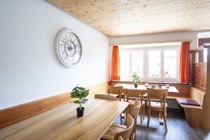 Uma área de refeições com mesa e cadeiras de madeira, um relógio na parede e uma janela com cortinas.