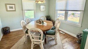Een eetkamer met een ronde houten tafel, zes stoelen, een hanglamp en twee ramen met jaloezieën.